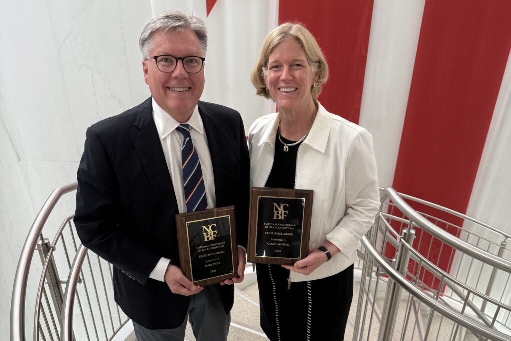 Mark, a white man with grey hair and black glasses, wears a black suit. McNeil, a white woman with blond hair, wears a black dress and white blazer. They stand