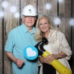 Gerard, a white man with grey hair, wears a teal polo, and Janet, a white woman with blond hair, wears a black blouse and khaki jacket. Janet holds an inflatable hammer, and Gerard is wearing a hard hat. 