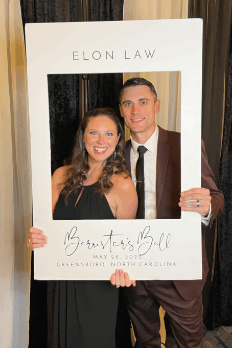 Bowyer, a white man with brown hair, wears a white shirt, brown tie and brown suit. His spouse is pictured with him. She is a white woman with brown hair who is wearing a black dress. They are holding a large white cutout frame that says "Barrister's Ball, May 28, 2022, Greensboro, NC."
