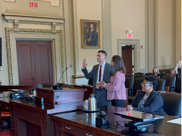 Bowyer, a white man with brown hair wearing a suit, stands at the podium with wife Jennifer. His hand is on the Bible. Keir Morton-Manley, a Black woman with black hair, is wearing a black and white dress and black blazer and is seated. 