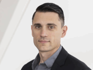 Bowyer, a white man with brown hair, wears a grey shirt and a black suit.