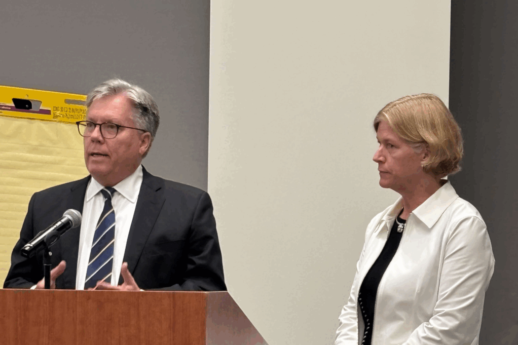Mark, a white man with grey hair and black glasses, wears a black suit, white shirt, and striped tie. McNeil, a white woman with blond hair, wears a black blouse and white jacket. Holt stands speaking at a podium.