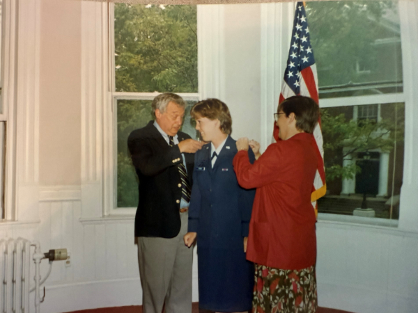Parady stands in the center in her military uniform, a navy jacket and skirt. Her father stands to her left and her mother to her right, and they are pinning on her military insignia. An American flag is visible in the background.