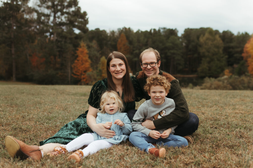 Ligon, a white woman, wears a dark green dress, and her husband, a white man, with brown hair and glasses, wears a dark green shirt and jeans. Her daughter, a little girl with blond hair, wears a blue dress, and her son, a boy with curly brown hair, wears a grey sweater. They are seated in a field.
