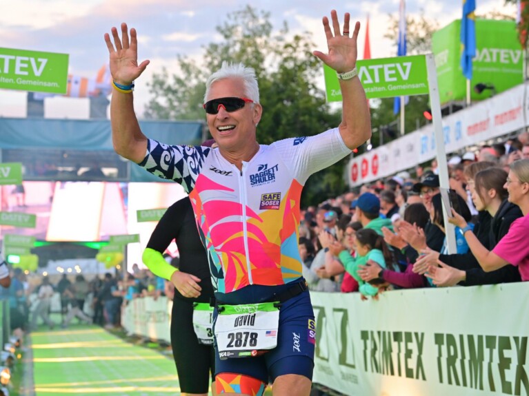 Daggett is wearing sunglasses and a white, navy and coral shirt and navy shorts as he crosses the finish line with his arms raised.
