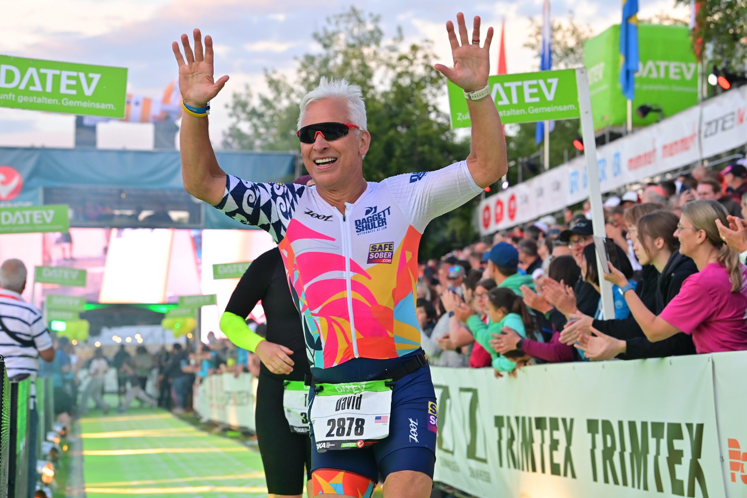 Daggett is wearing sunglasses and a white, navy and coral shirt and navy shorts as he crosses the finish line with his arms raised.