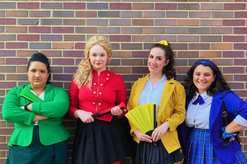 Risser wears a green blazer and is crossing her arms, Alyssa wears a red blazer and is holding a fan, Autumn wears a yellow blazer and is holding a yellow fan, and Sid wears a blue blazer and is smiling.
