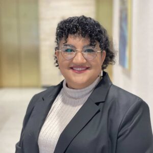 Hannah, a woman with curly dark brown hair, wears glasses, a grey sweater and dark grey blazer.