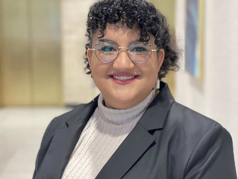 Hannah, a woman with curly dark brown hair, wears glasses, a grey sweater and dark grey blazer.