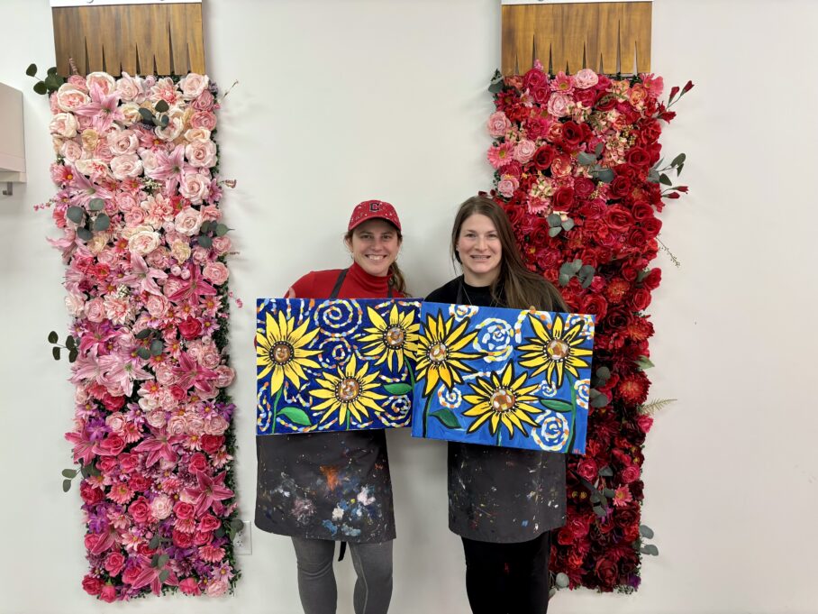Ligon, a white woman with brown hair, and Erin, a white woman with brown hair and wearing a red hat, are smiling and each holding painted canvases with sunflowers.
