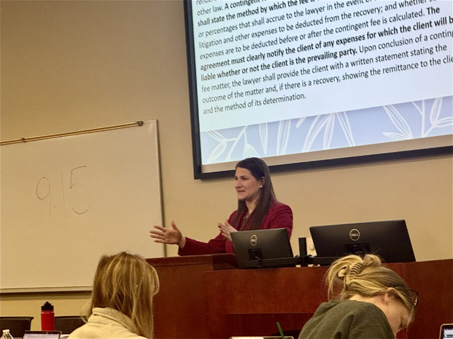 Ligon, a white woman with brown hair, wears a red suit and is teaching students at a podium. students are pictured taking notes.