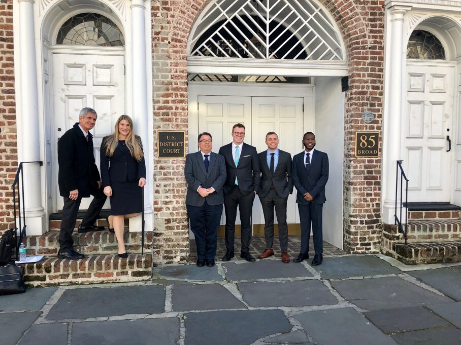 Ligon wears a black dress, and all other individuals are pictured wearing suits. They stand in front of the U.S. District Court in Charleston, a brown brick building with white doors.