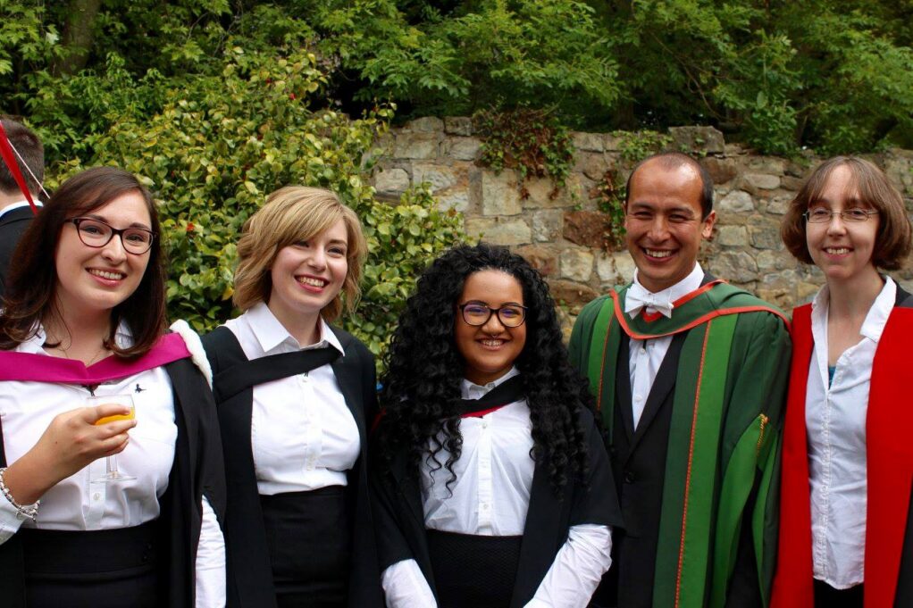 Risser, a woman with black hair and glasses, is photographed in a white shirt, black skirt and black gown. Her professors are pictured in white shirts with green and red college gowns, and her friends are pictured in white shirts with black college gowns.