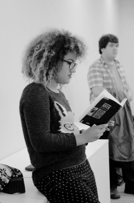 Risser is photographed in black and white and wearing glasses, a dark sweater and polka dotted pants. She is reading the play and a young man is standing in the background.