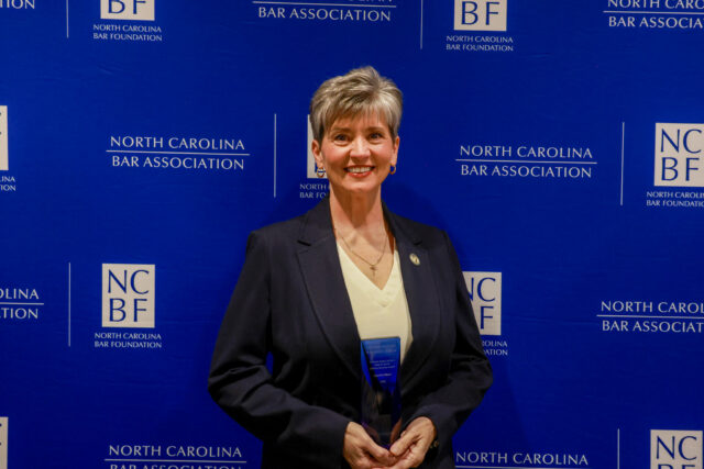 Cynthia, a white woman with grey hair, wears a white blouse and black blazer. She is holding the award, which is blue.