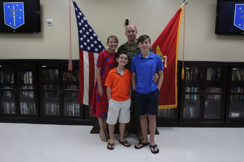Plummer wears his uniform, and he is standing with his two sons, one wearing an orange shirt and the other wearing a blue shirt, and wife Hannah, who is wearing a red dress. He stands with the American flag and Marine Corps flag behind him.