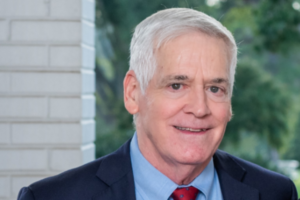 Gray, a white man with white hair, wears a blue shirt, red tie and dark grey jacket.