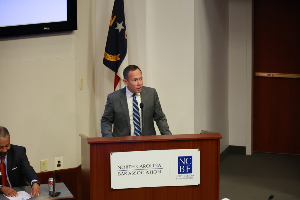 Jason, a white man with brown hair, wears a white shirt, blue and white striped tie and grey suit. He is speaking at a podium with the NC flag behind him.
