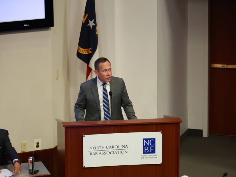 Jason, a white man with brown hair, wears a white shirt, blue and white striped tie and grey suit. He is speaking at a podium with the NC flag behind him.