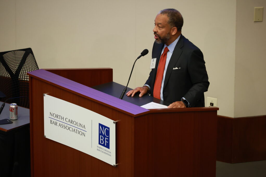Rob, a Black man with black hair and a beard, wears a blue shirt, red tie and black suit and stands at a podium.