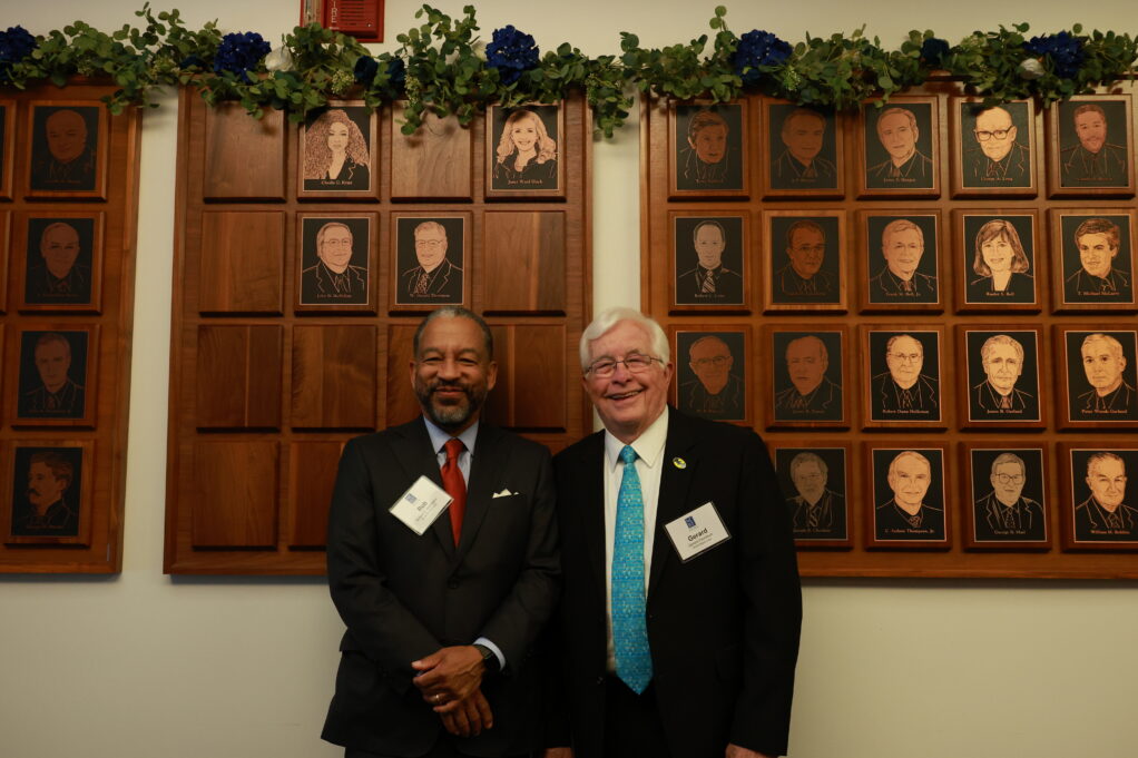Rob, a Black man with black hair and a beard, wears a blue shirt, red tie and black suit. Gerard, a white man with white hair, wears a white shirt, teal tie and black suit.