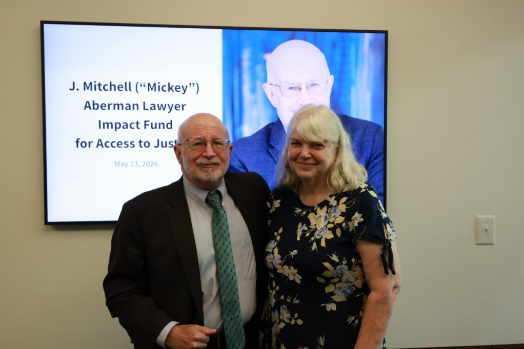 Mickey, a white man with a shaved head and a white beard, wears a blue shirt, teal tie and black suit. His spouse, a white woman with grey hair, wears a black dress with white flowers.