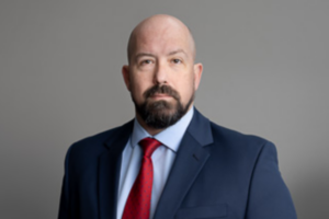 Plummer, a white man with a shaved head and a brown beard and mustache, wears a blue shirt, red tie and navy suit.
