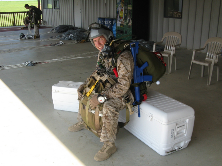 Plummer, wearing his military uniform and gear for the training jump, rests on a white ice chest and is smiling.