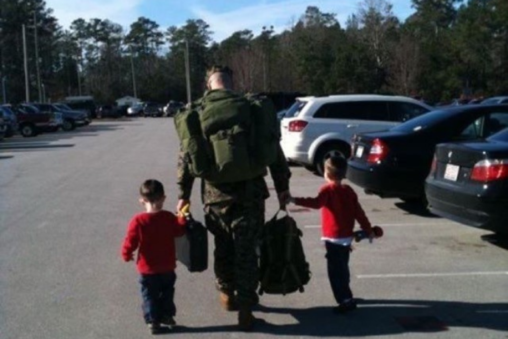 Plummer, a white man with brown hair, holds the hand of each of his sons, who are toddlers and are walking on either side of him. He is in his military uniform and each son is wearing a red shirt. They are walking with their backs to the camera in a parking lot.