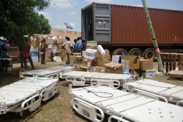 A group of Black individuals are photographed near a large truck. There are many boxes and medical supplies, including gurneys, photographed in the foreground.