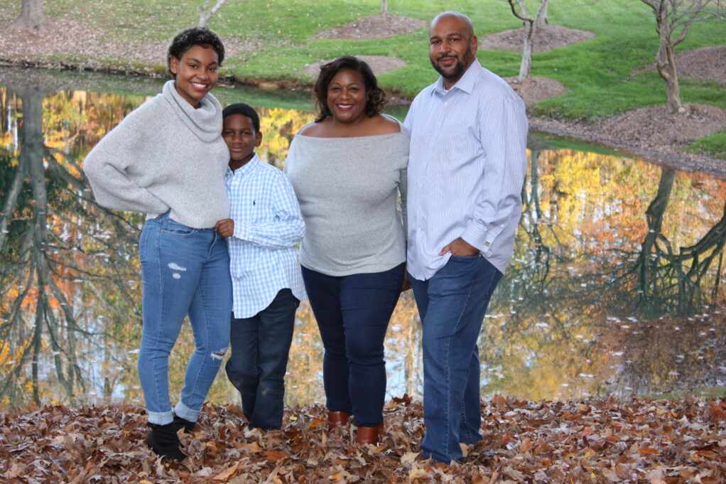 Micah a Black woman with brown hair, wears a grey blouse. Her husband, a Black man with a beard, wears a blue shirt. Her daughter wears a grey sweater and her son wears a blue shirt. They are standing outside.