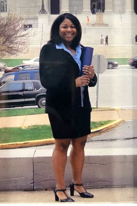 Micah, a Black woman with black hair, wears a blue shirt and black suit. She is standing outside.