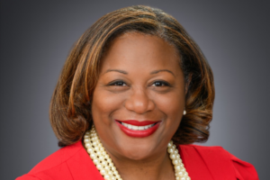 Micah, a Black woman with brown hair, wears a red suit and a pearl necklace.