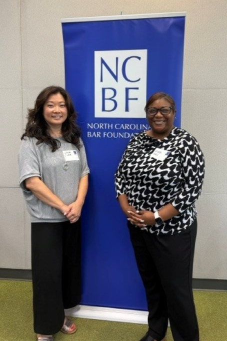 Micah, a Black woman with brown hair, wears a black and white blouse. She is photographed with another volunteer, and they are standing in front of the NCBF logo sign.