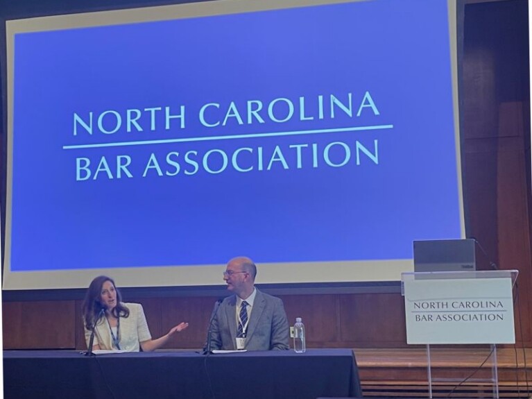 Lauren, a white woman with brown hair, wears a cream colored blouse, and Rob wears a white shirt, black tie and grey suit. They are seated at a table in front of the room.