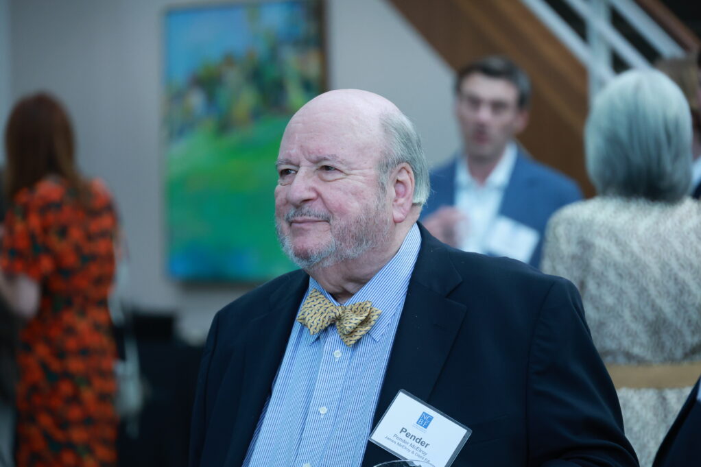 Pender, a white man with grey hair, wears a blue shirt, yellow bowtie and black suit.