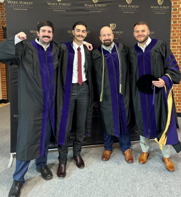 Plummer is dressed in his law school doctoral gown and is standing with three fellow law school graduates who are also wearing law school doctoral gowns and smiling.