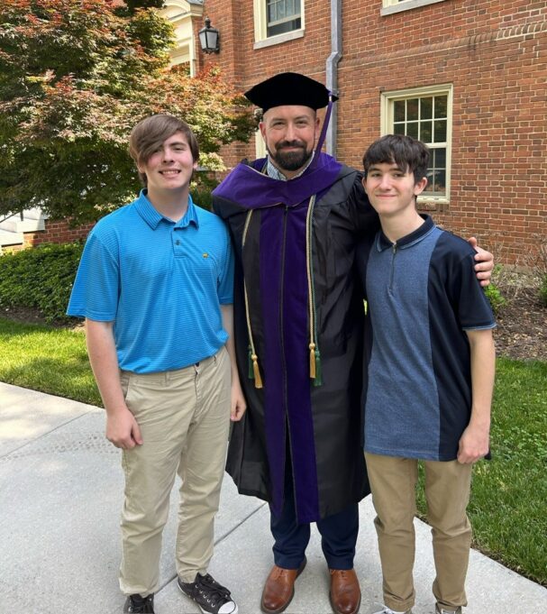 Plummer is wearing his law school doctoral gown and tam and stands with one son on either side. They are photographed outside in front of a brick building.