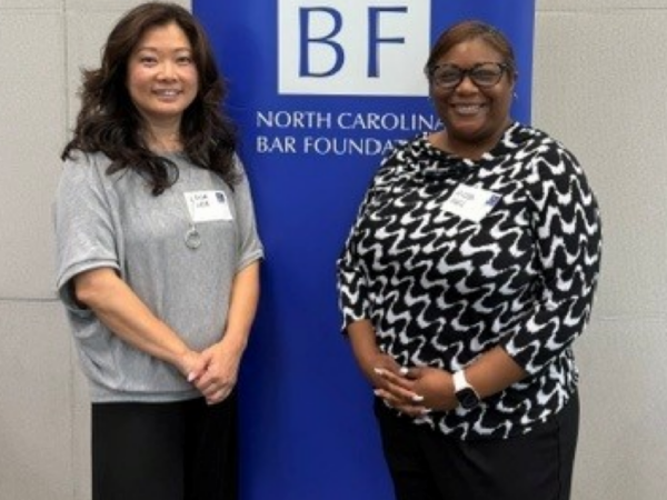 Micah, a Black woman with Black hair, wears a blouse with a black and white pattern. She stands with a woman who has black hair and is wearing a grey blouse. An NCBF sign is standing behind them.