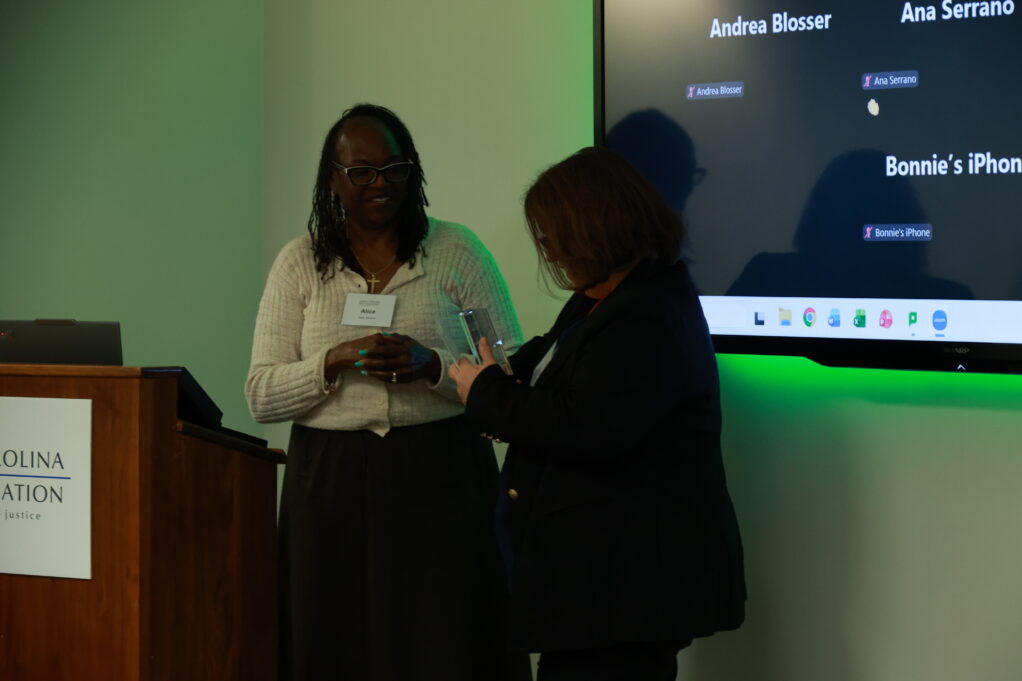 Alice, a Black woman with black hair, wears a cream colored sweater and stands at a podium. 