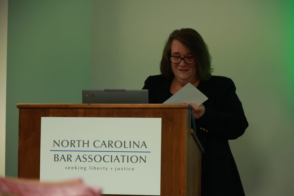 Smith, a white woman with brown hair and glasses wears light blouse and black blazer. She is standing at a podium. 