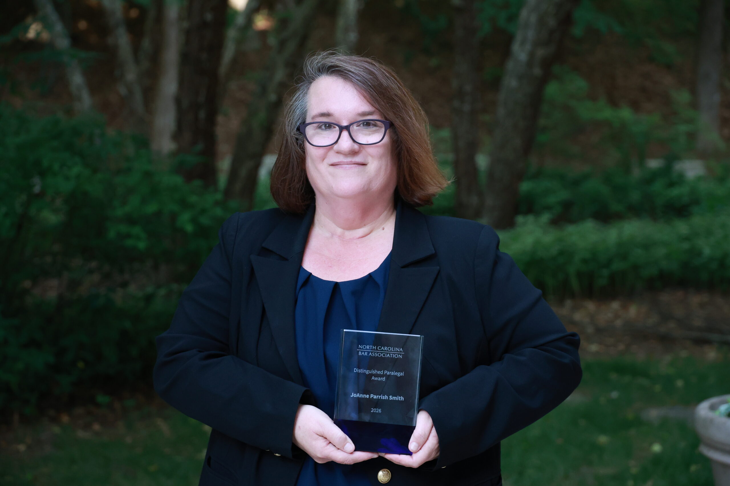 JoAnne, a white woman with brown hair, wears a blue blouse and black blazer. She is standing outside and holding her award. 