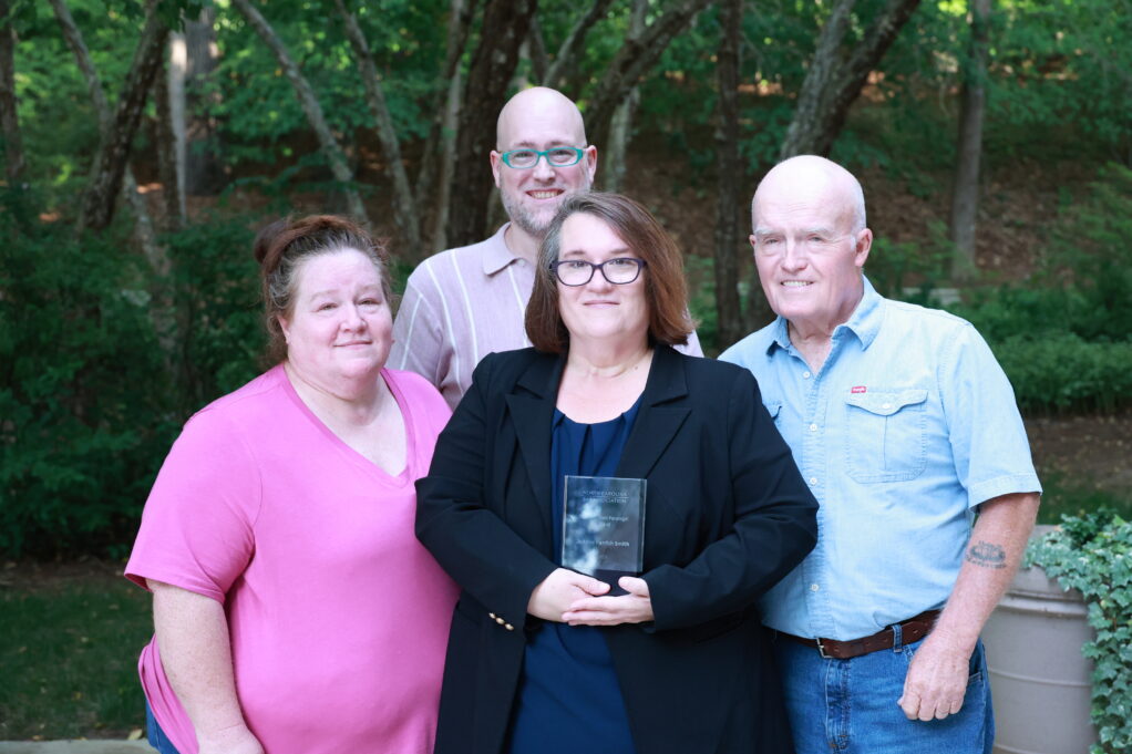 Smith, a white woman with brown hair, is wearing a blue blouse and black blazer. She is pictured holding the award. Her sister, a white woman with brown hair stands to her right, her brother, a white man with a shaved head and glasses, stands behind her, and her father, a white man with grey hair, stands to her left. 